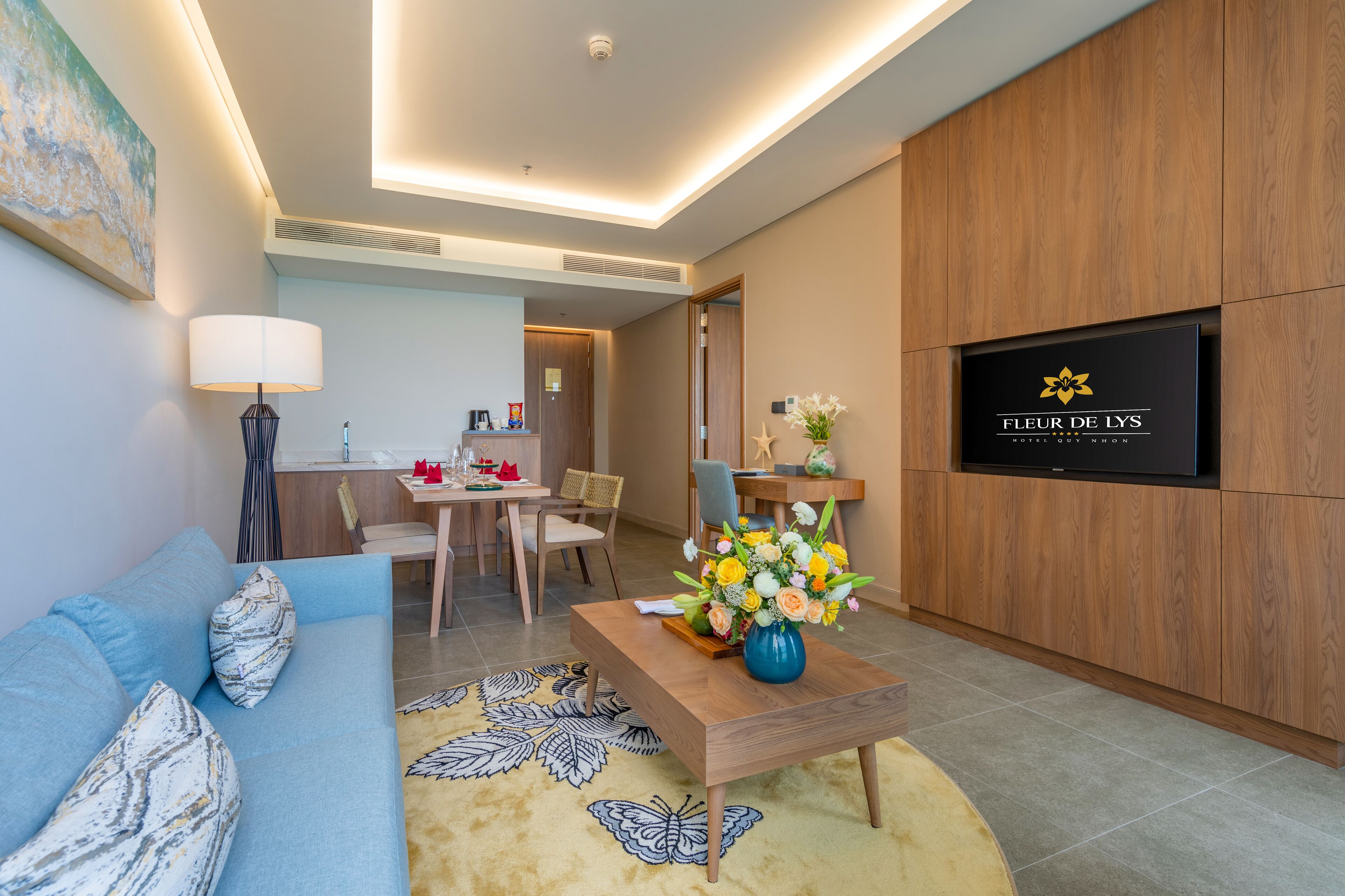 A modern hotel room with a sofa, dining table, and a TV mounted on the wall.