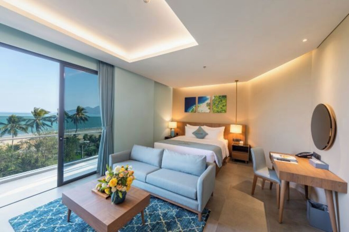 Ein modernes Hotelzimmer mit Bett, Sofa, Schreibtisch und einem großen Fenster mit Blick auf Palmen und Berge.