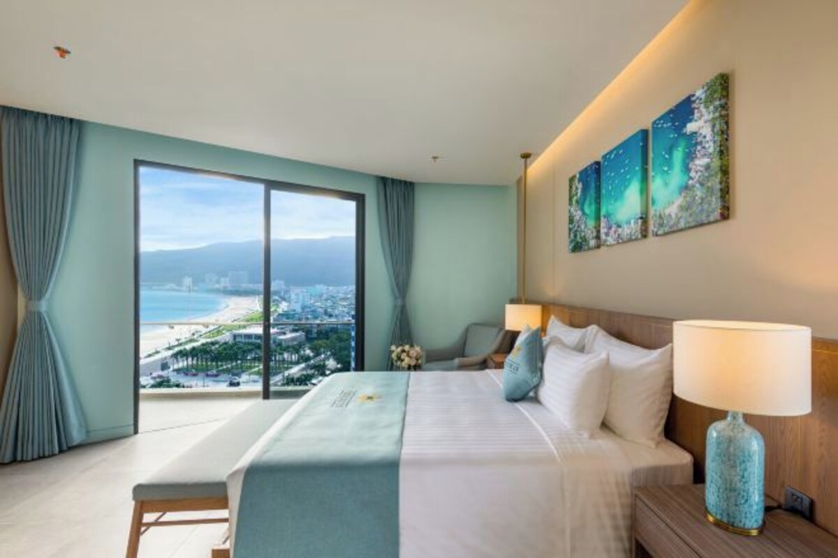 Een hotelkamer met een groot bed, een nachtkastje, een lamp en uitzicht op de stad en het strand door een groot raam.