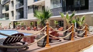 Ein Poolbereich mit Liegestühlen und Palmen, umgeben von einer Holzterrasse und einem Gebäude mit mehreren Balkonen.