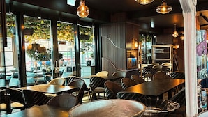 Ein Restaurantinneres mit mehreren Tischen und Stühlen, hängenden Pendelleuchten und großen Fenstern mit Blick auf die Straße.