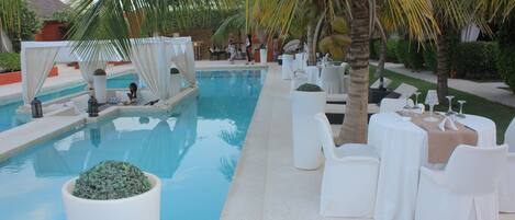 A poolside area with a clear pool, white lounge chairs, and tables set for dining.