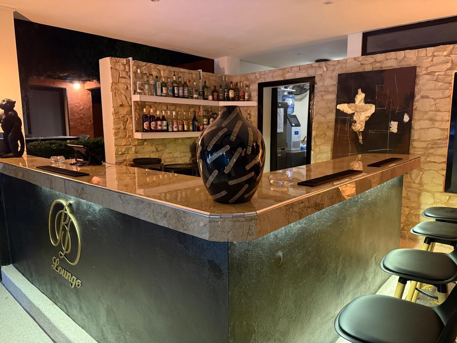 Uma área de bar com parede de pedra, prateleiras com garrafas de bebidas, um vaso decorativo grande e um logotipo no balcão.