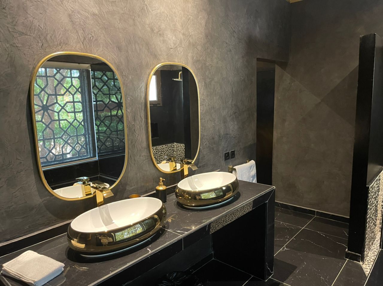 Un baño con dos lavamanos, cada uno con un grifo dorado y un espejo grande arriba. Las paredes tienen una textura decorativa, y hay una ventana con un diseño geométrico.