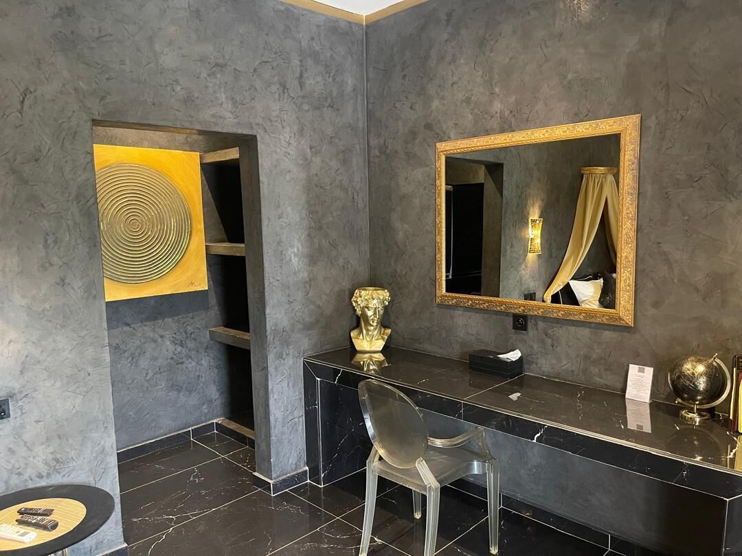 A modern bathroom with a dark marble vanity, a gold-framed mirror, and a clear chair.