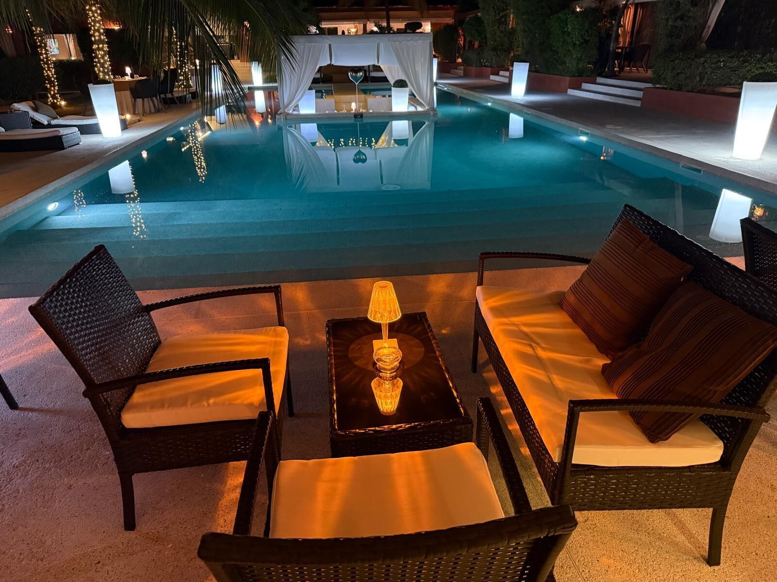 A poolside seating area with wicker chairs and a table, surrounded by a pool with lights reflecting on the water.