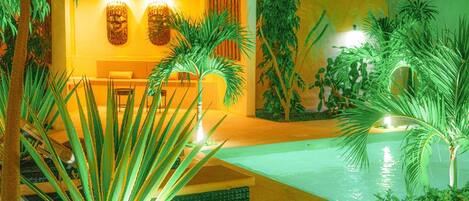 Un espacio exterior moderno con piscina, rodeado de plantas tropicales y una zona de estar con mesa y sillas.