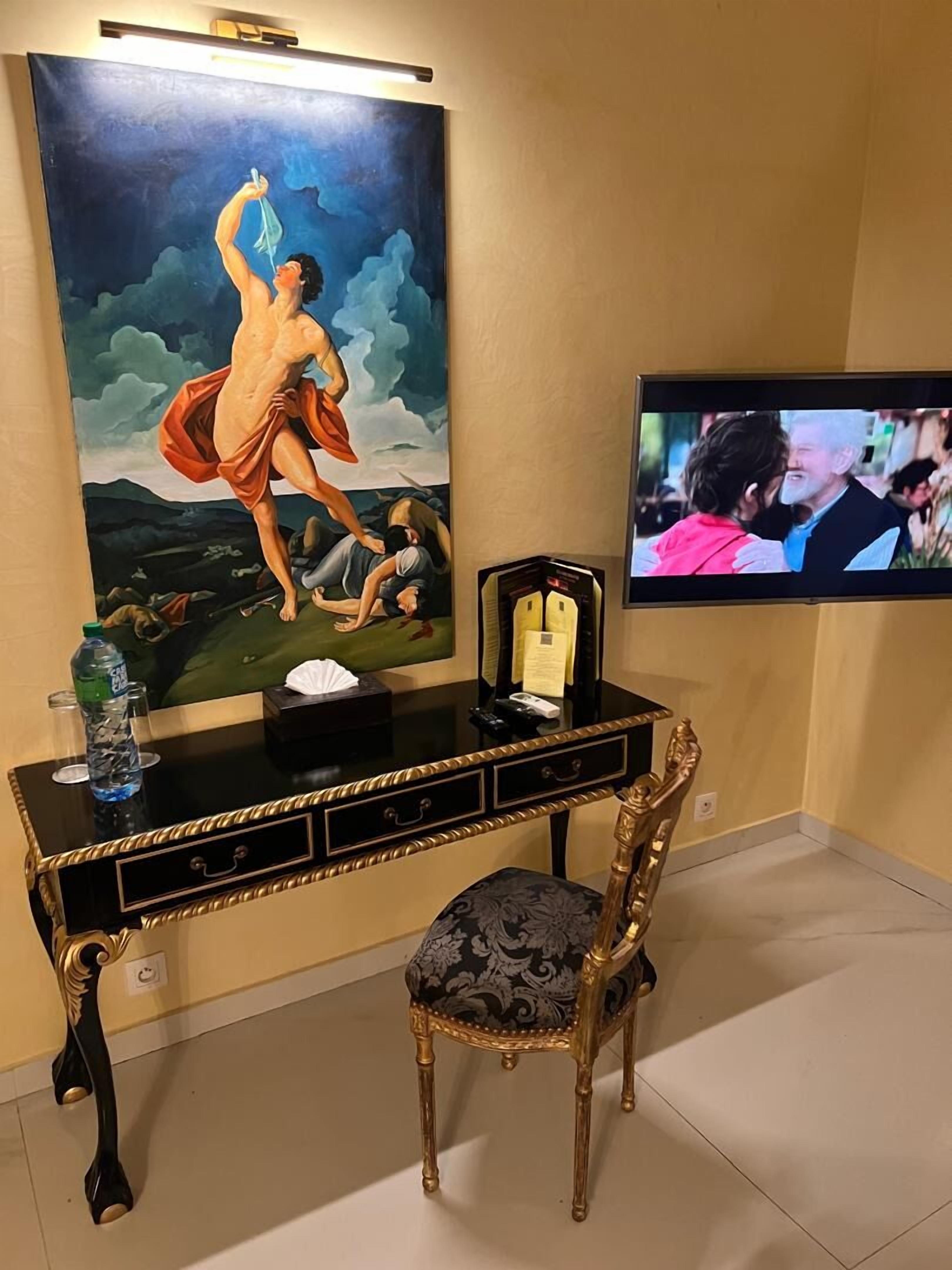 A room with a large painting, a television, a desk with drawers, a chair, and a bottle of water.