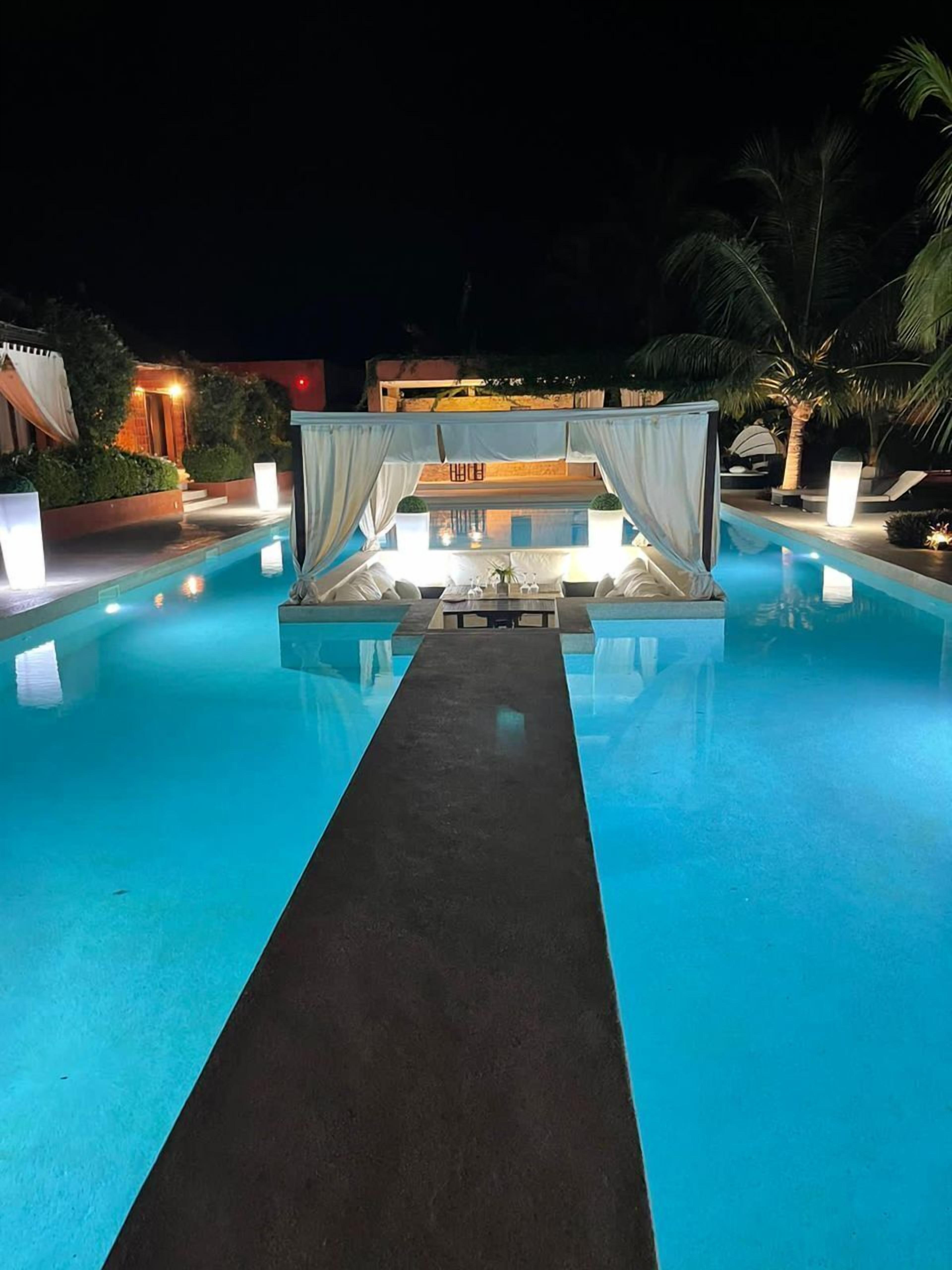 A nighttime pool area with a central seating area, palm trees, and ambient lighting.