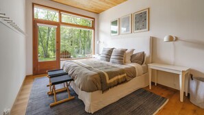 Ein Schlafzimmer mit einem großen Bett, Holzboden, einer Bank, einem Tisch und Blick auf Bäume durch ein großes Fenster.