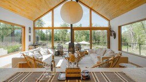 A spacious living area with a high ceiling, large windows, and a central fireplace. - A Retreat @ Hudson Woods With Pool & Hot Tub (Kerhonkson)