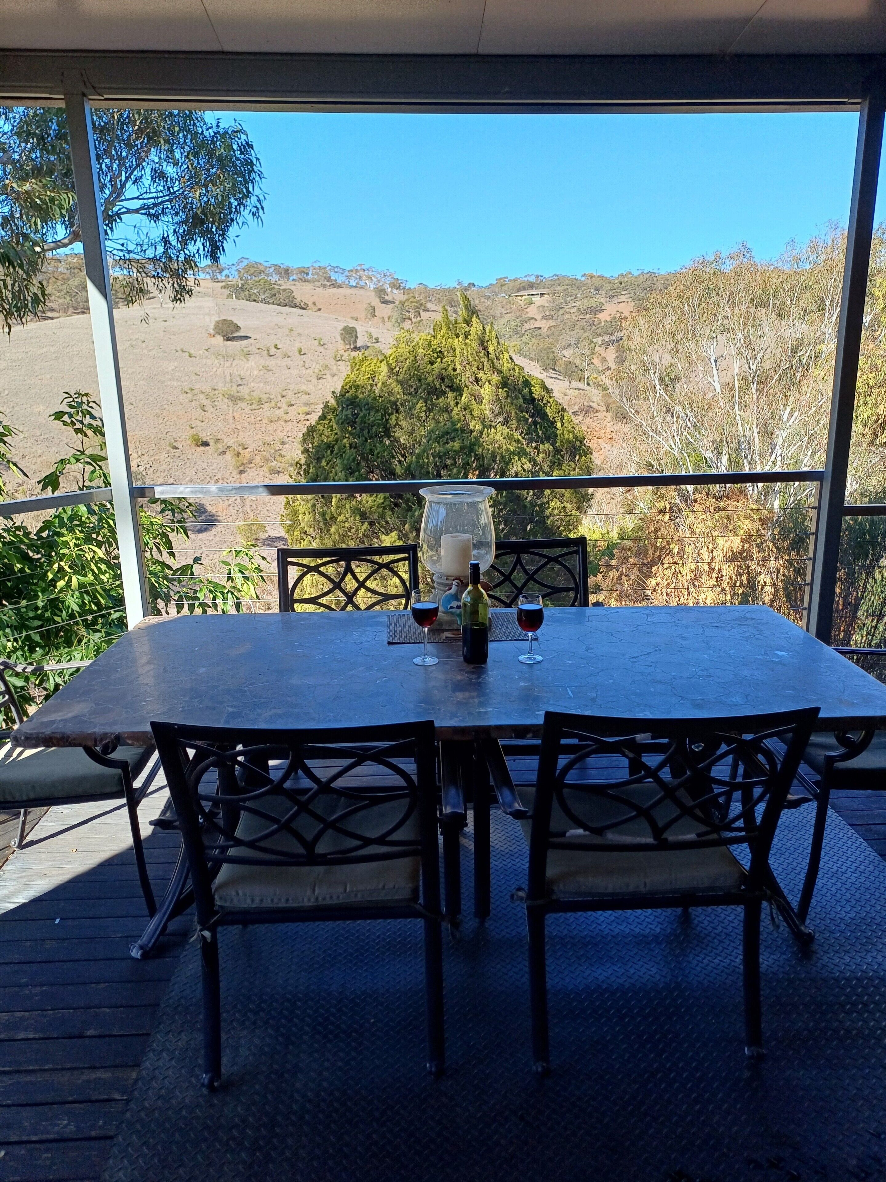 An outdoor dining area with a table and chairs, a glass of wine, and a view of a hilly landscape.