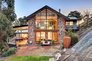 Una casa de piedra de dos plantas con ventanas amplias, un patio con mobiliario exterior y un camino de piedra que lleva a la entrada.