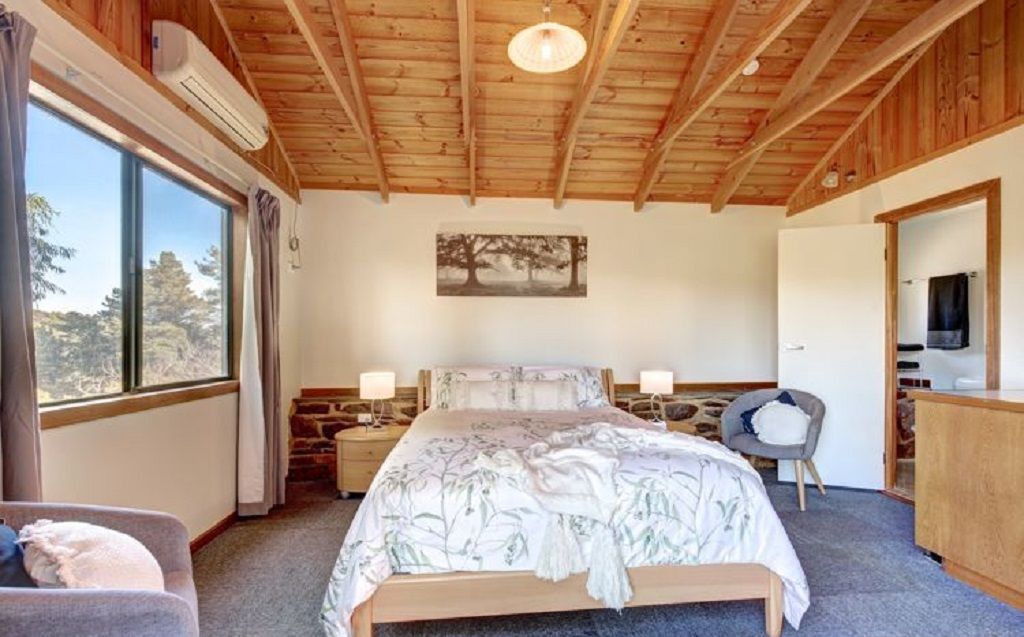A bedroom with a wooden ceiling, a large bed with a floral bedspread, a nightstand with a lamp, a chair, and a view of trees through a window.