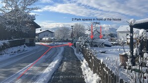 A snowy residential street with a house on the left, two park spaces in front, and a clear sky.