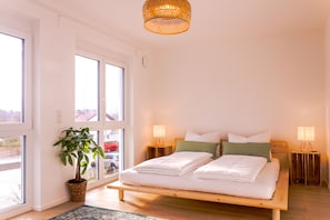 Ein Schlafzimmer mit einem Bett, Nachttischlampen, einer Topfpflanze und großen Fenstern.