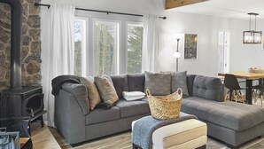 A cozy living room with a stone wall, a fireplace, a grey sofa, a wooden dining table, and a large window with white curtains. - Great views from condo w/free winter shuttle (Brian Head)