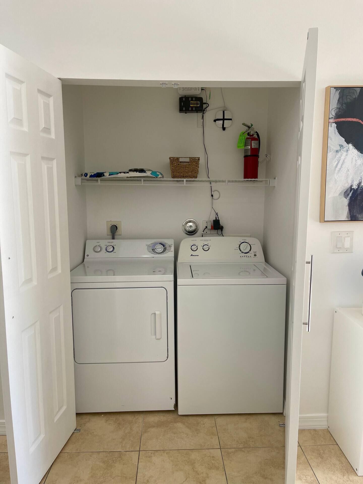 A laundry room with two white washing machines, a fire extinguisher, and a shelf with items.