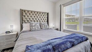 Een slaapkamer met een gestuukt hoofdeinde, een bed met grijze beddengoed, een blauwe plaid, nachtkastjes met lampen en een raam met zonwering.