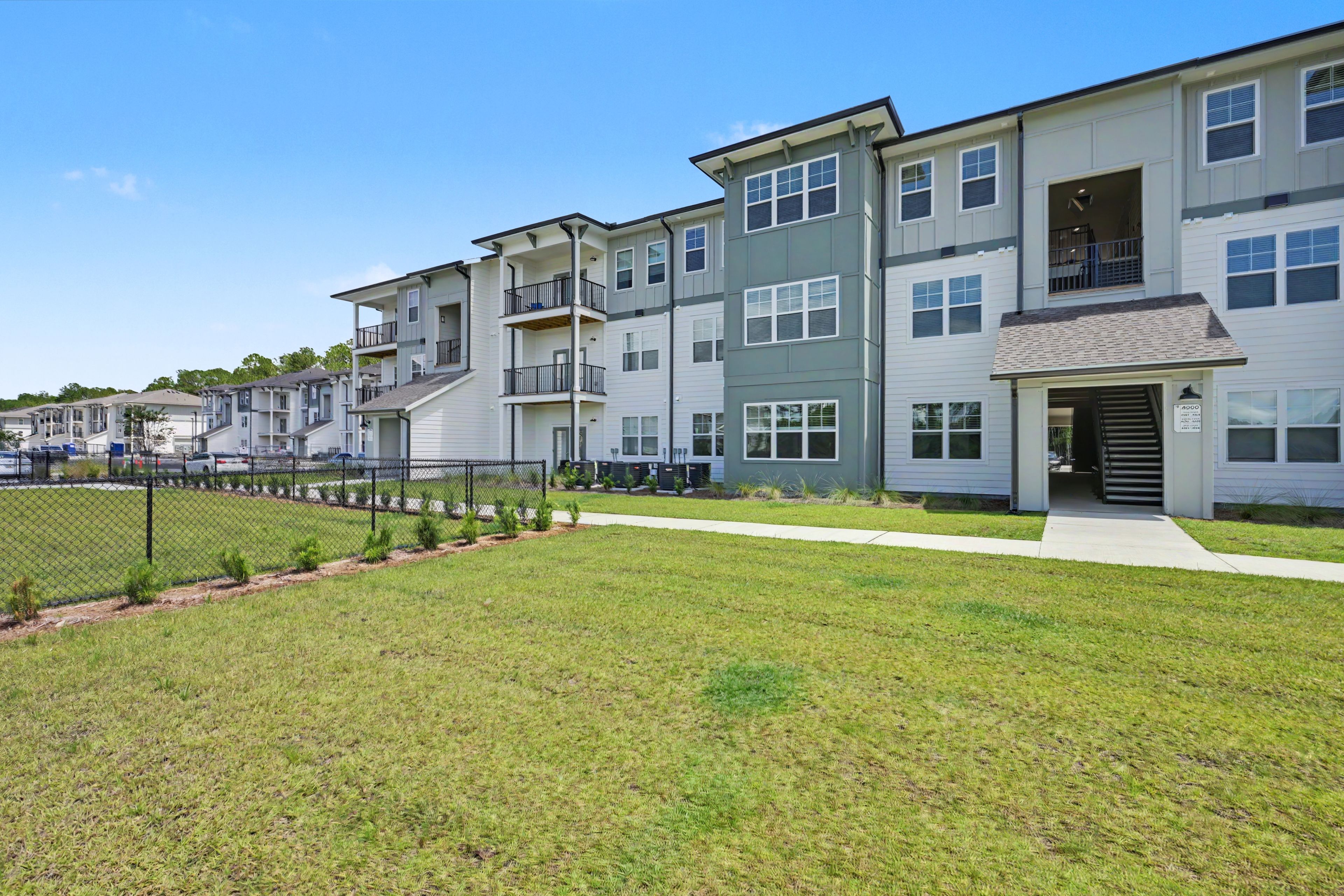 A row of modern multi-story apartment buildings with balconies, a fenced parking area, and a well-maintained lawn.