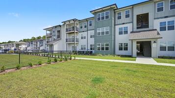 A row of modern multi-story apartment buildings with balconies, a fenced parking area, and a well-maintained lawn.