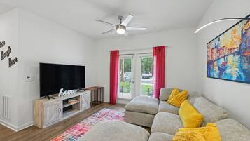 A living room with a flat-screen TV, a ceiling fan, a sofa with yellow cushions, and a colorful rug.