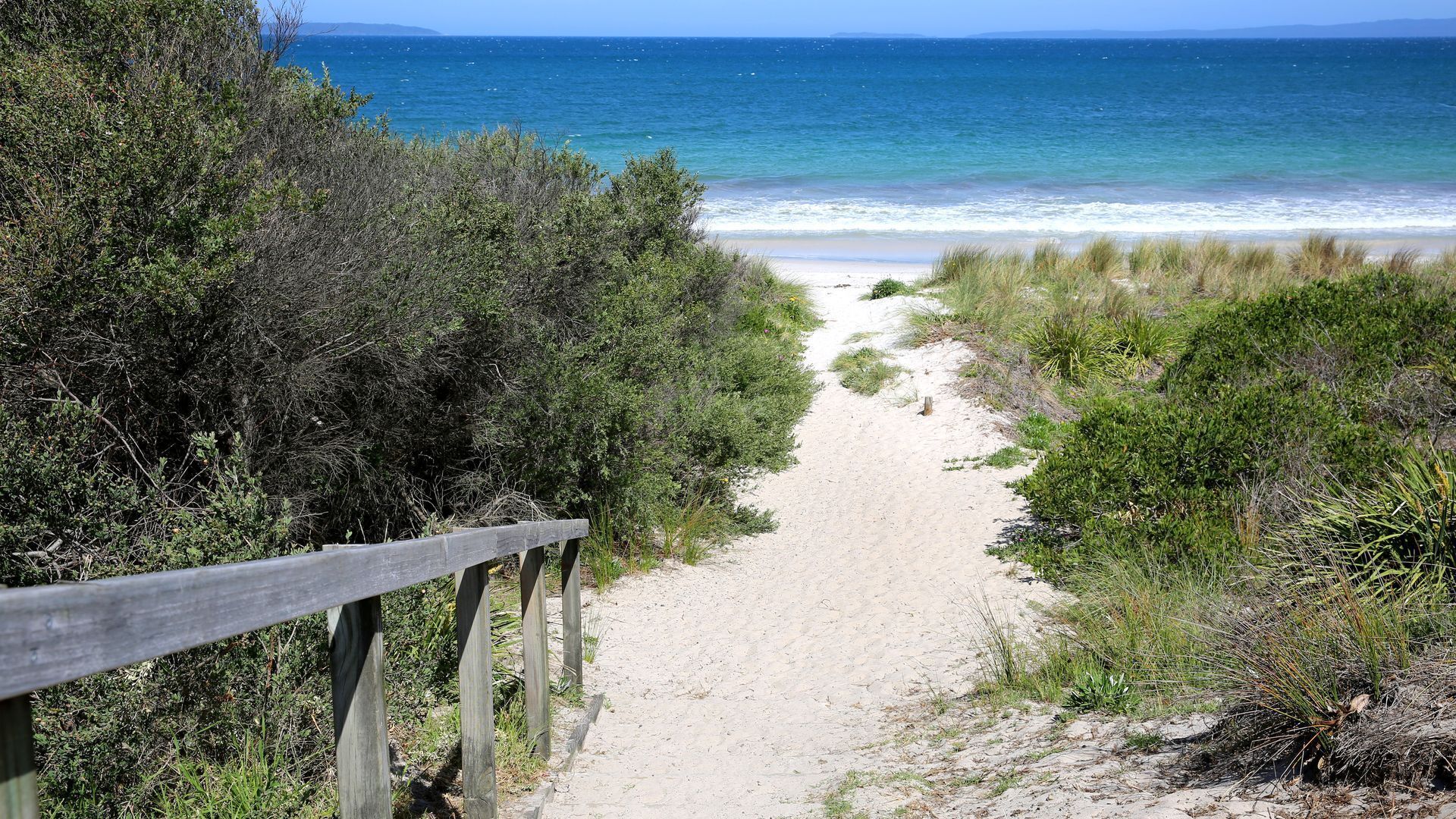 A sandy beach path with a wooden fence on one side, leading to the ocean.