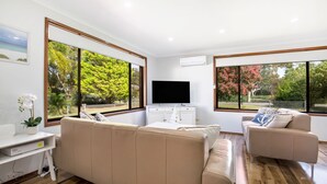 Ein modernes Wohnzimmer mit einem Flachbildfernseher, beigen Sofas und großen Fenstern mit Blick auf Bäume und einen Park.