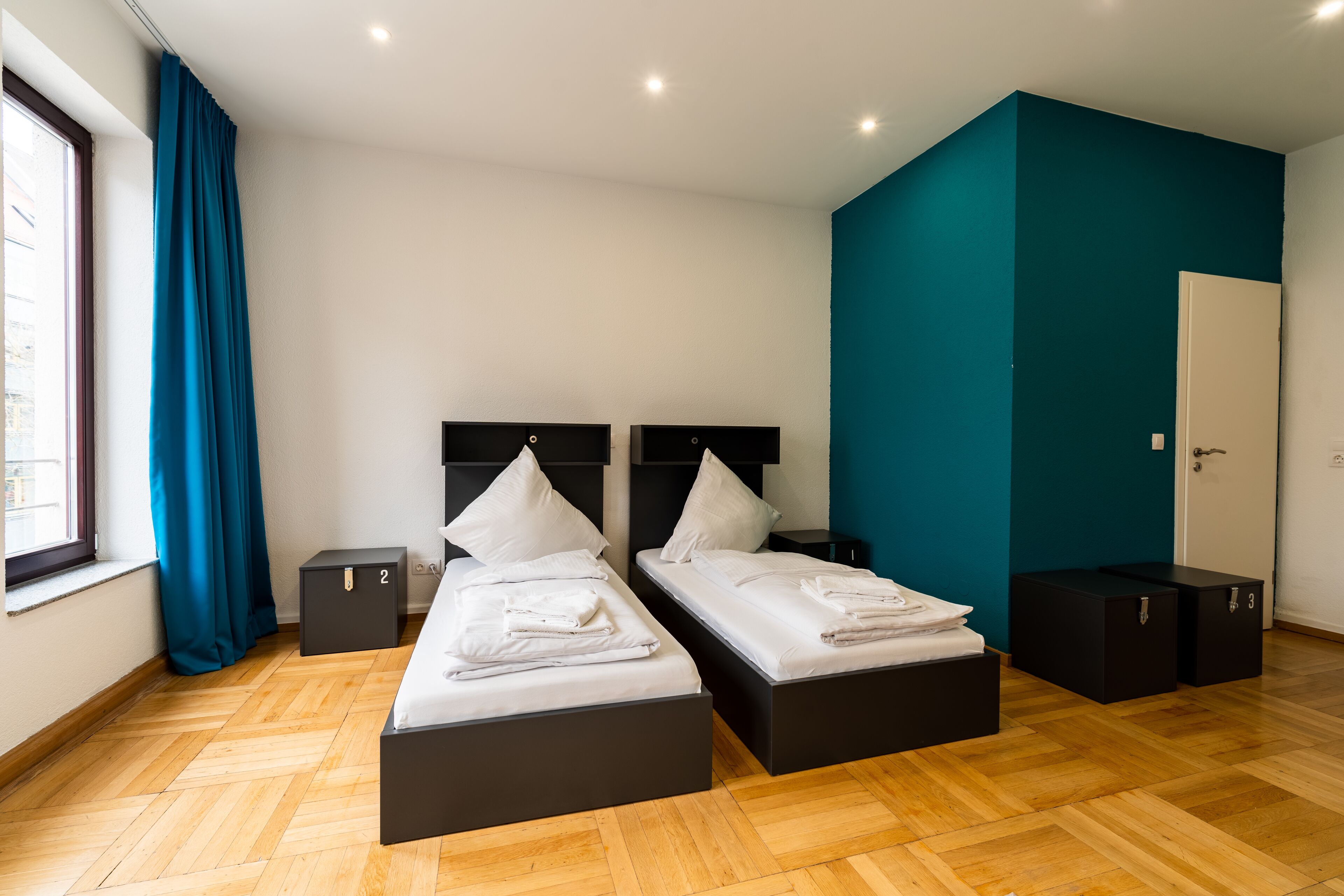 Una habitación de hotel moderna con dos camas, una pared de acento color turquesa y un amplio ventanal con cortinas.