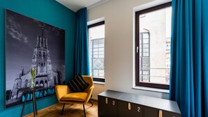 A room with a large black-and-white photo of a cathedral, a yellow chair, a black cabinet with drawers, and a window with blue curtains.