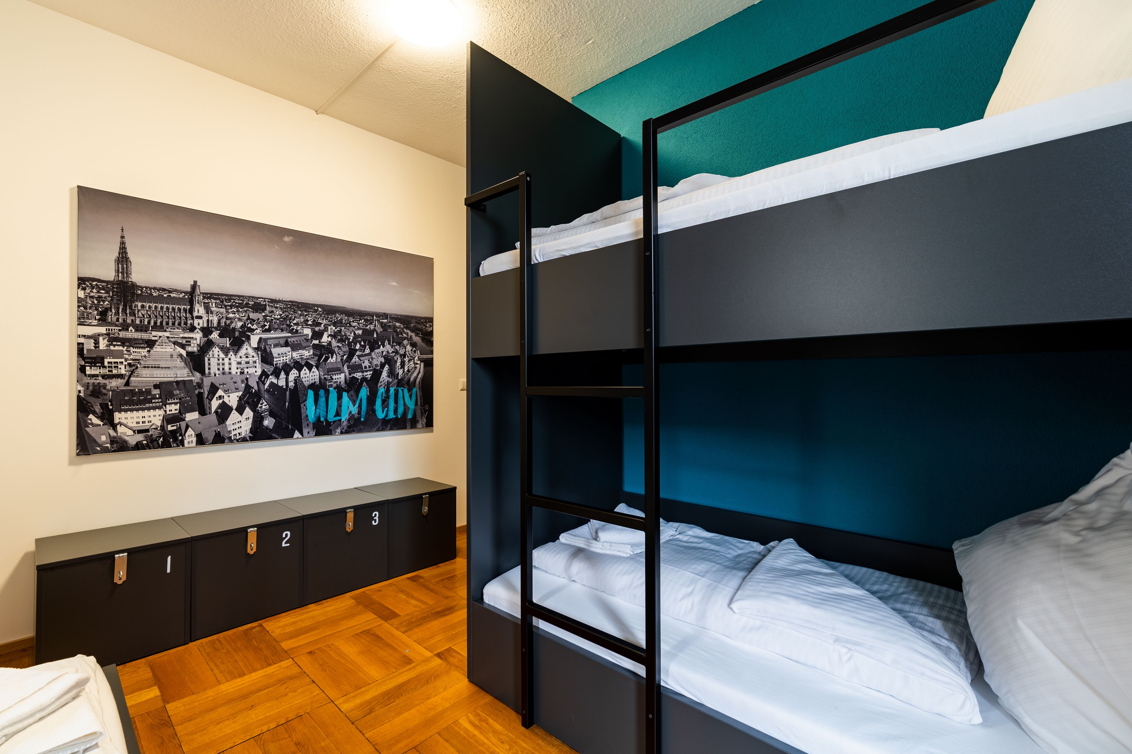 Una cama litera con ropa de cama blanca, piso de madera, un mueble de almacenamiento fijado a la pared con compartimentos numerados y un cartel con vista a la ciudad.