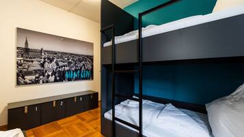 Una cama litera con ropa de cama blanca, piso de madera, un mueble de almacenamiento fijado a la pared con compartimentos numerados y un cartel con vista a la ciudad.