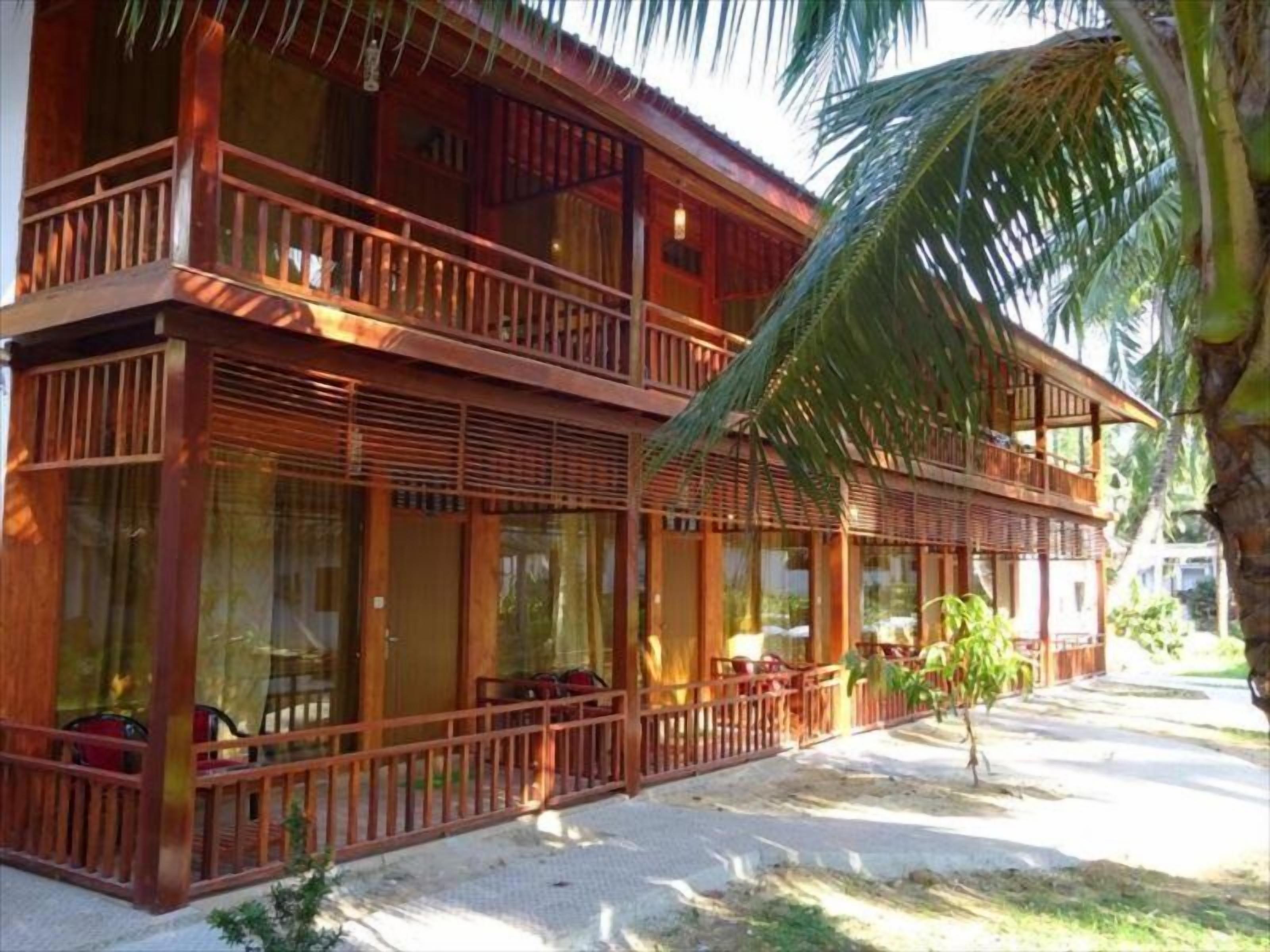 A two-story wooden building with balconies, large windows, and a palm tree in the foreground.