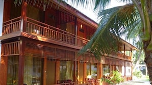 A two-story wooden building with balconies, large windows, and a palm tree in the foreground.