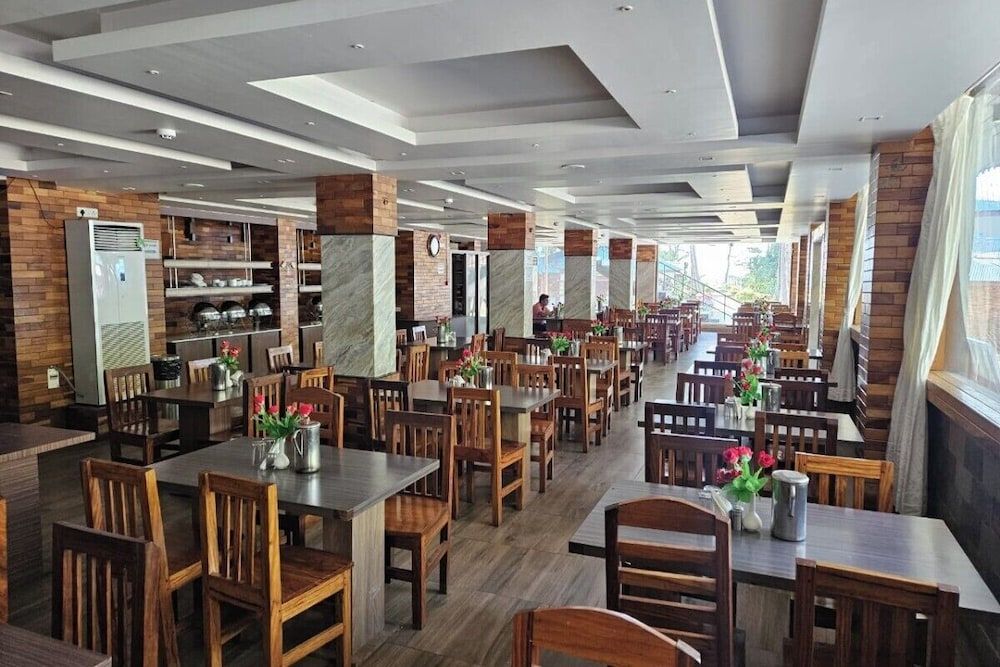 A spacious restaurant with wooden tables and chairs, a central serving area, and large windows along one side.