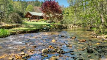 A cabin by a clear stream with a wooden deck and a large tree with red leaves.