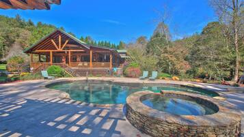 A lodge with a wooden exterior, a stone hot tub, and a pool area surrounded by trees.
