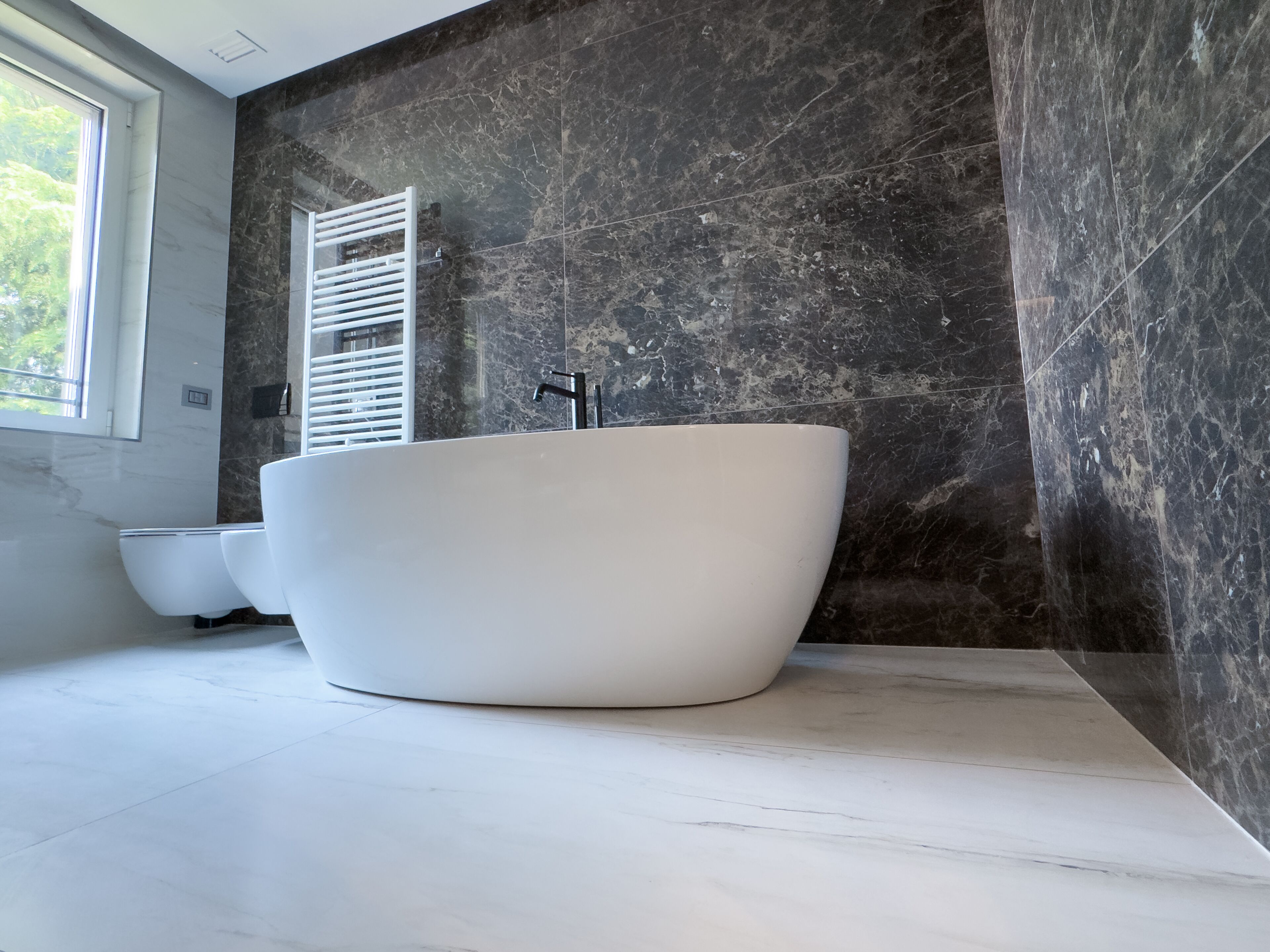 A modern bathroom with a freestanding bathtub, a window with blinds, and a marble wall.
