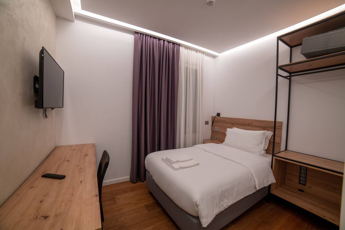 A modern hotel room with a wooden desk, a bed with white linens, a TV mounted on the wall, and a built-in shelving unit.