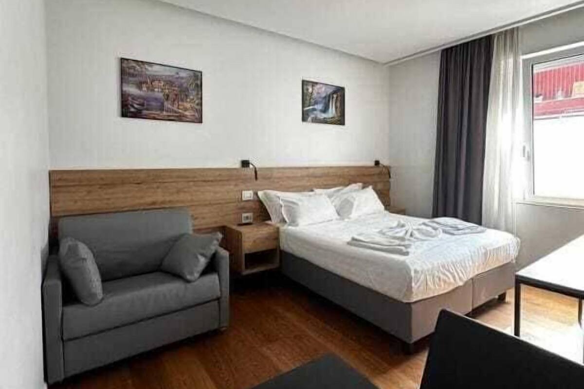 A modern hotel room with a bed, a grey sofa, a small table, and two paintings on the wall.