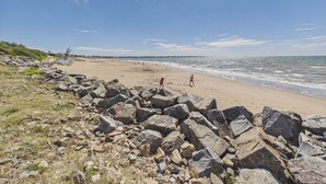Ein Strand mit großen Felsen im Vordergrund, einem sandigen Ufer und dem Meer im Hintergrund.