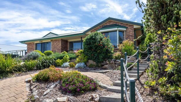 A two-story house with a brick exterior, a green roof, and a landscaped garden with various plants and flowers. - Dive into Dorset - 10 Dorset Court (CARRICKALINGA)
