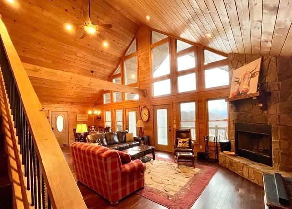 A spacious living room with a fireplace, wooden ceiling, large windows, and a staircase.