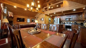 A spacious dining area with a high ceiling, wooden beams, and a chandelier. A kitchen with modern appliances is visible in the background. - Sunset Ridge Lodge - Sunset Ridge Lodge - Breathtaking mountain views and (Bryson City)