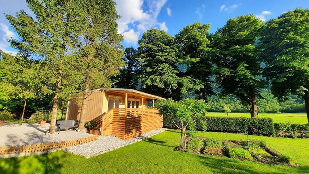 Eine Holzhütte mit Veranda und kleinem Garten, umgeben von Bäumen und einer Wiese.