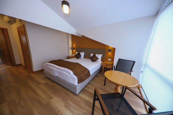 A modern hotel room with a large bed, a small round table, and a chair. There is a wooden floor and a ceiling light fixture.