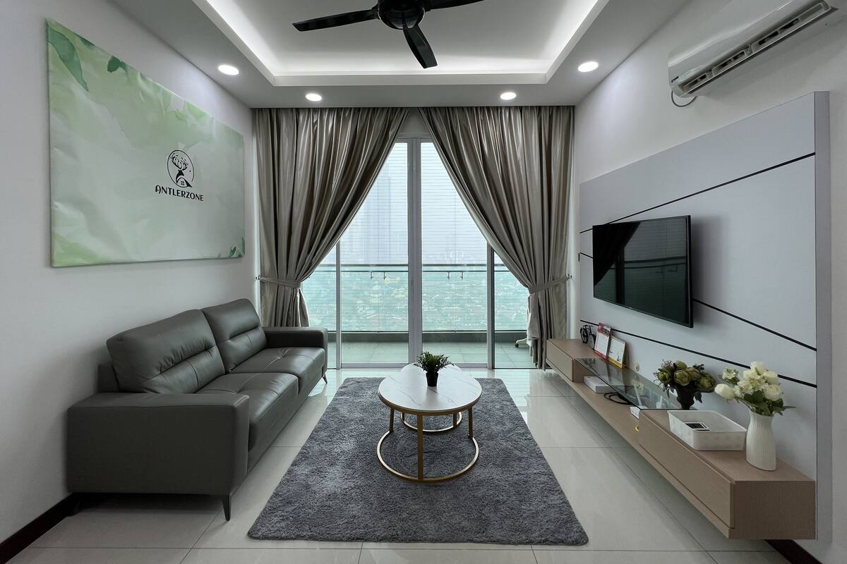 A modern living room with a grey sofa, a round coffee table, a ceiling fan, a TV mounted on the wall, and a large window with curtains.
