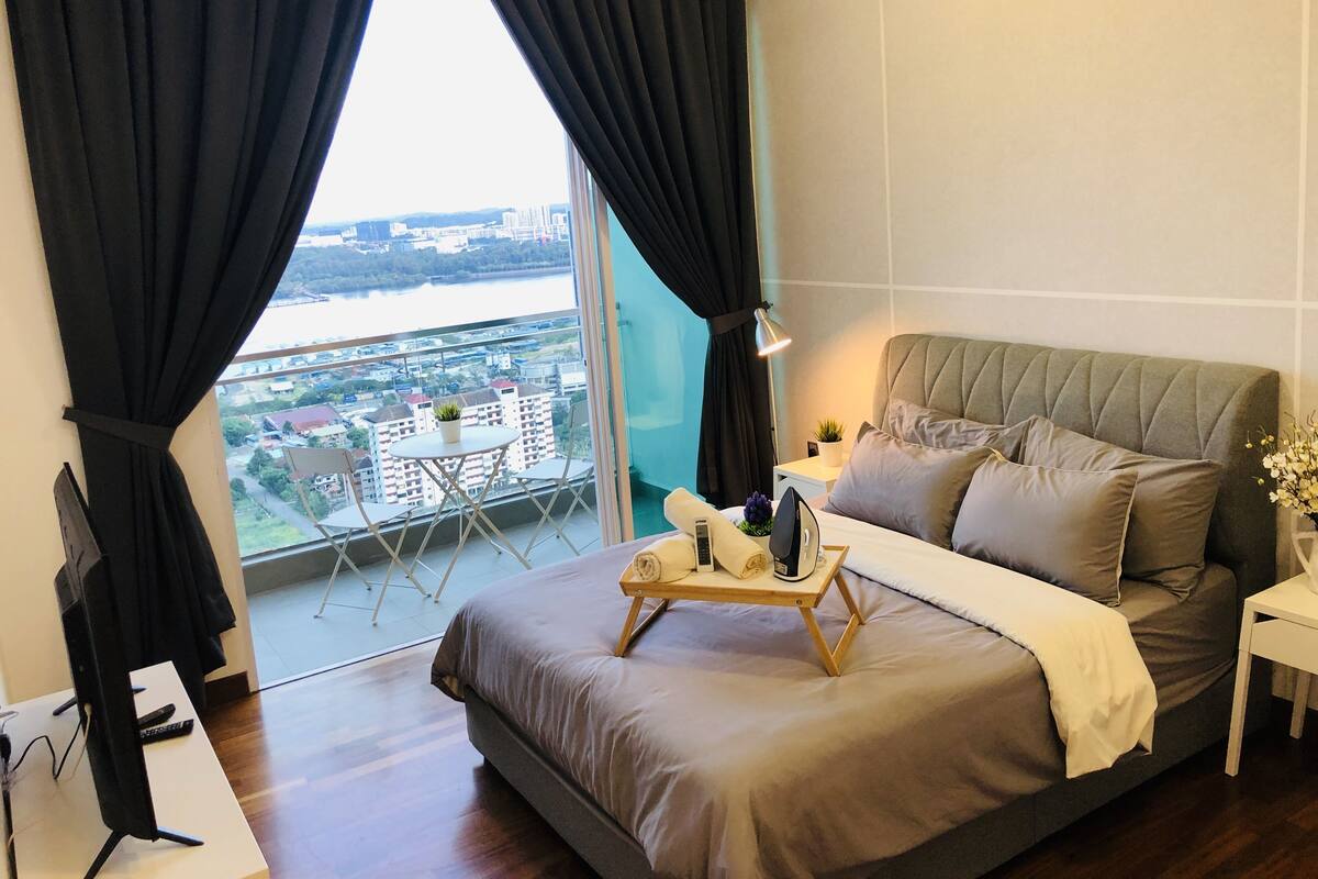 A bedroom with a large bed, a small table with a lamp, a TV, and a view of a cityscape through a window.