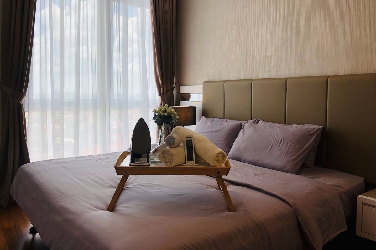 A neatly made bed with a tray holding an iron, a hairdryer, and a towel, a bedside table with a lamp, and sheer curtains allowing natural light.
