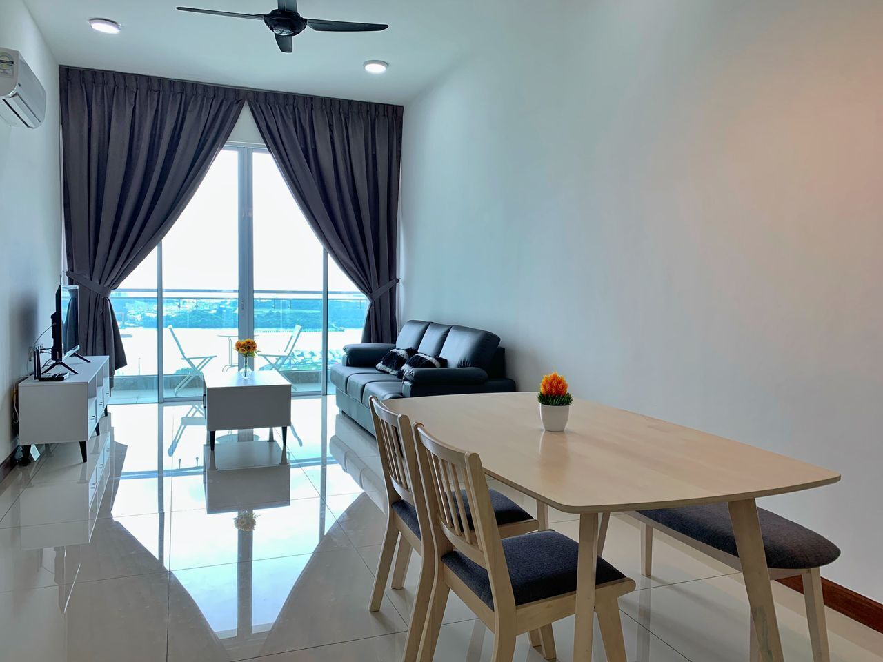 Un salon moderne avec un canapé, une table à manger et un téléviseur. Une grande fenêtre, ornée de rideaux, laisse entrevoir un balcon.